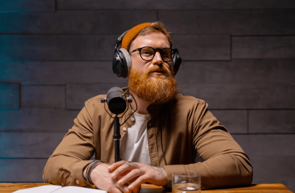 Mężczyzna z rudą brodą i w czapce nagrywa podcast, siedząc przed mikrofonem z nałożonymi słuchawkami, w tle ściana z ciemnych cegieł