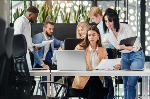 Zespół zróżnicowanych pracowników współpracuje w nowoczesnym biurze typu open space. Kobieta siedzi przy laptopie, analizując dokumenty, a inni współpracownicy stoją i dyskutują wokół niej.