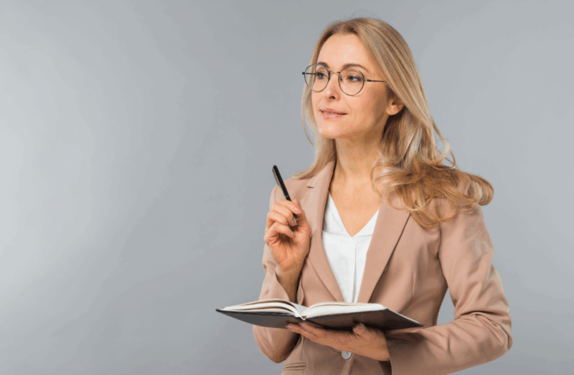 Kobieta w okularach i beżowej marynarce, trzymająca otwarty notatnik i długopis, zamyślona spogląda w bok