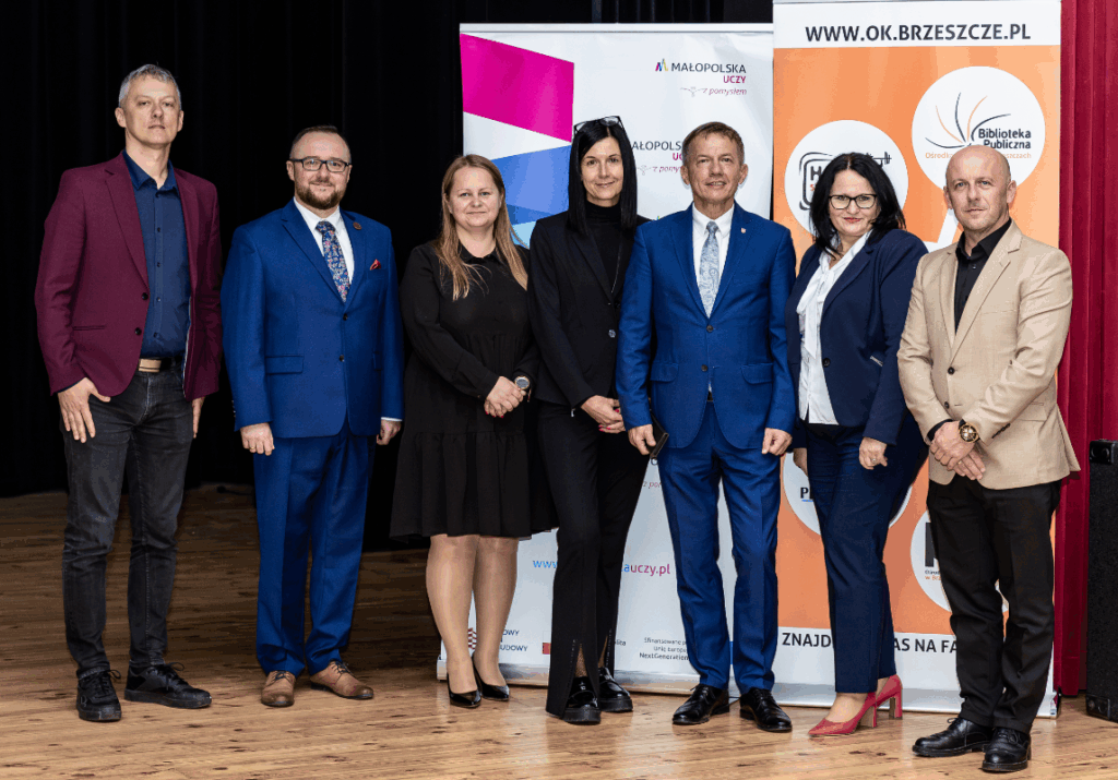 Grupa osób w eleganckich strojach pozuje do zdjęcia. W tle roll up Małopolska Uczy i Ośrodka Kultury w Brzeszczach.