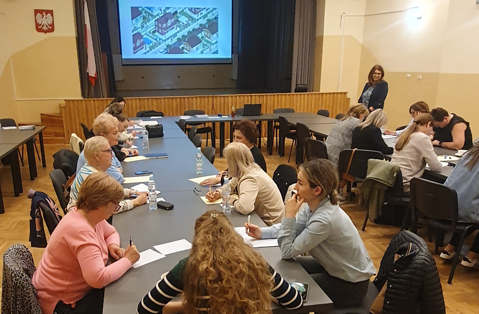 Kobiety w różnym wieku siedzą przy dwóch długich stołach, w dużej sali z drewnianym parkietem, uczestnicząc w warsztatach. W tle, na podwyższeniu, na ekranie projektora widoczna jest prezentacja, a obok ekranu stoi kobieta (prelegentka). W sali widoczna jest również flaga Polski.