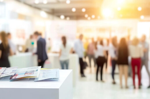 Widok z perspektywy stoiska informacyjnego na tętniące życiem targi w nowoczesnym wnętrzu. Na pierwszym planie widać białą ladę z rozłożonymi ulotkami i broszurami informacyjnymi.