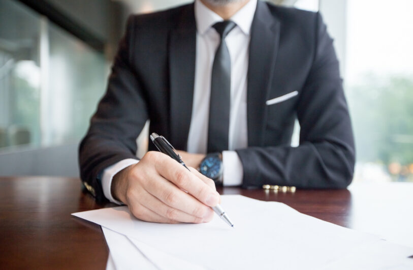 Mężczyzna w eleganckim ciemnym garniturze i krawacie podpisuje piórem dokumenty leżące na biurku. Postać w tle jest rozmyta, co kieruje uwagę na dokumenty.
