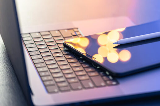 Zbliżenie na klawiaturę laptopa, na której ukośnie leży tablet z ekranem odbijającym rozmyte żółte okręgi światła i nowoczesny rysik ułożony na jego prawym brzegu. Kompozycja utrzymana jest w chłodnej kolorystyce z ciepłymi, rozmytymi odblaskami światła na ekranie.