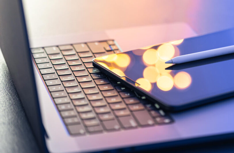 Zbliżenie na klawiaturę laptopa, na której ukośnie leży tablet z ekranem odbijającym rozmyte żółte okręgi światła i nowoczesny rysik ułożony na jego prawym brzegu. Kompozycja utrzymana jest w chłodnej kolorystyce z ciepłymi, rozmytymi odblaskami światła na ekranie.