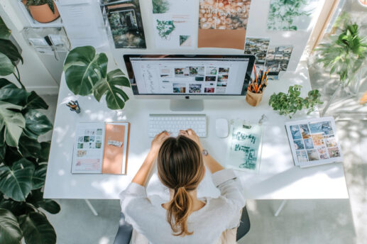 Widok z góry na jasne, białe biurko, przy którym siedzi kobieta, jej dłonie spoczywają na białej klawiaturze przed dużym monitorem wyświetlającym miniatury zdjęć; na blacie leżą notesy i dokumenty, mysz, kubek z długopisami oraz kilka doniczek z roślinami, a za monitorem na ścianie wisi kolaż fotografii i ilustracji.