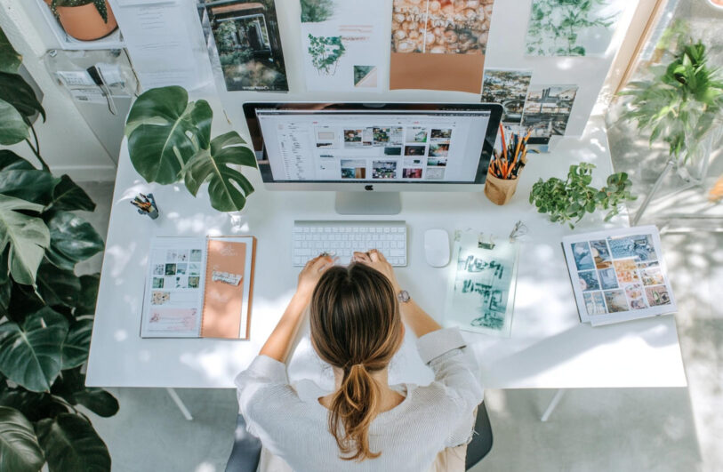 Widok z góry na jasne, białe biurko, przy którym siedzi kobieta, jej dłonie spoczywają na białej klawiaturze przed dużym monitorem wyświetlającym miniatury zdjęć; na blacie leżą notesy i dokumenty, mysz, kubek z długopisami oraz kilka doniczek z roślinami, a za monitorem na ścianie wisi kolaż fotografii i ilustracji.