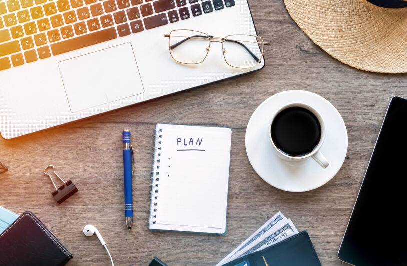 Widok z góry na drewniany blat biurka: w lewym górnym rogu częściowo widoczny laptop z okularami na klawiaturze, na środku notatnik z napisem „PLAN” i niebieski długopis obok, po prawej filiżanka kawy na spodku oraz telefon.