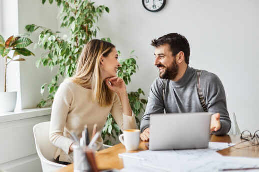 Dwoje dorosłych siedzi przy drewnianym stole — kobieta w kremowym swetrze i mężczyzna z brodą w szarym swetrze z szelkami — patrzą na siebie i uśmiechają się, na stole widoczny laptop, filiżanka kawy, rozłożone dokumenty i okulary, w tle duże doniczkowe rośliny oraz zegar na ścianie.