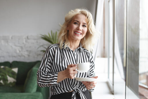 Kobieta z blond włosami uśmiecha się stojąc przy dużym oknie, ubrana w czarno-białą koszulę w pionowe paski i trzyma białą filiżankę na spodku, w tle widoczna zielona sofa i roślina.
