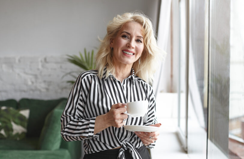 Kobieta z blond włosami uśmiecha się stojąc przy dużym oknie, ubrana w czarno-białą koszulę w pionowe paski i trzyma białą filiżankę na spodku, w tle widoczna zielona sofa i roślina.