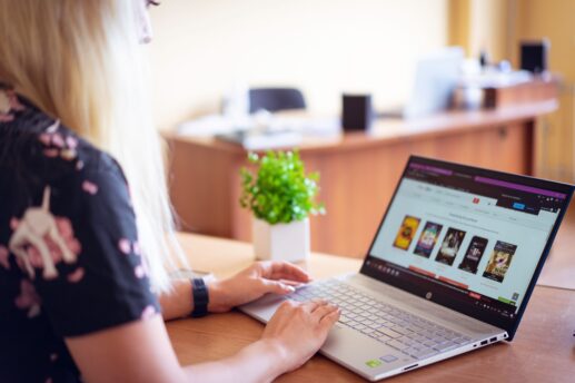 Osoba z długimi blond włosami siedzi przy drewnianym biurku i używa srebrnego laptopa z widocznymi miniaturami filmów na ekranie, obok stoi mała zielona roślina w białej doniczce, a w tle widać rozmyte biurowe meble.