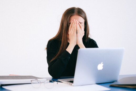 Kobieta z długimi brązowymi włosami siedzi przy biurku za srebrnym laptopem Apple, zakrywając dłońmi twarz; na stole leżą okulary i rozłożone dokumenty, a tło jest jasne.