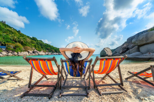 Na plaży trzy drewniane leżaki w jaskrawe, wielokolorowe paski; środkowy zajmuje osoba odwrócona tyłem w dużym białym kapeluszu z czarną wstążką, z rękami splecionymi za głową i długimi ciemnymi włosami, na pierwszym planie jasny, kamienisty piasek, dalej turkusowe morze otoczone po lewej zielonym, zalesionym wzgórzem z palmami, a po prawej dużymi płaskimi głazami pod błękitnym niebem z białymi chmurami.