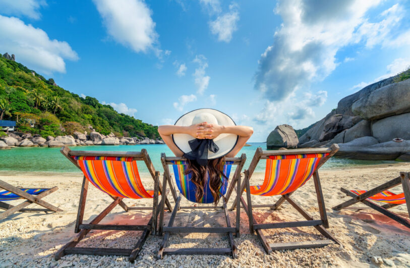 Na plaży trzy drewniane leżaki w jaskrawe, wielokolorowe paski; środkowy zajmuje osoba odwrócona tyłem w dużym białym kapeluszu z czarną wstążką, z rękami splecionymi za głową i długimi ciemnymi włosami, na pierwszym planie jasny, kamienisty piasek, dalej turkusowe morze otoczone po lewej zielonym, zalesionym wzgórzem z palmami, a po prawej dużymi płaskimi głazami pod błękitnym niebem z białymi chmurami.