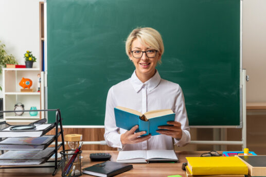 Kobieta siedzi za biurkiem pod dużą zieloną tablicą w klasie, trzyma otwartą niebieską książkę; ma krótkie blond włosy, czarne okulary i białą koszulę, a na stole przed nią leżą notatnik, segregator z dokumentami, koszyk z długopisami, kalkulator i żółte pudełko.